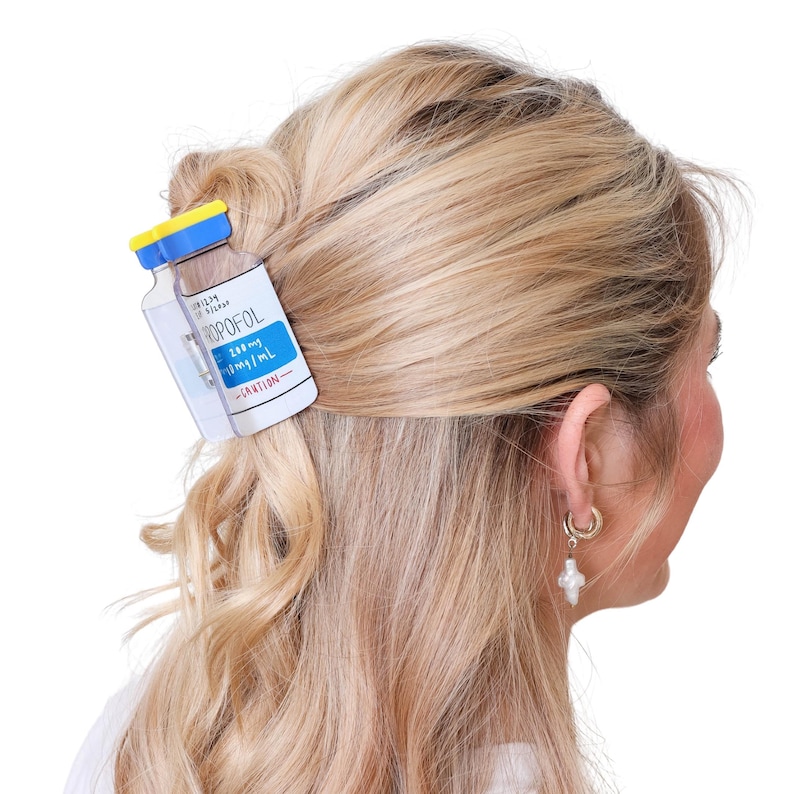May include: A hair claw clip shaped like a medical vial with a yellow cap and a white label featuring the text PROPOFOL, 200mg/mL, and CAUTION. The clip is secured in blonde hair, holding a section of hair back with its functional gripping mechanism.