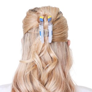 May include: A hair claw clip featuring a design that mimics a prescription medication bottle. The clip is clear with blue and white labels, yellow top accents, and a central metal spring mechanism, securing a section of blonde hair in place.