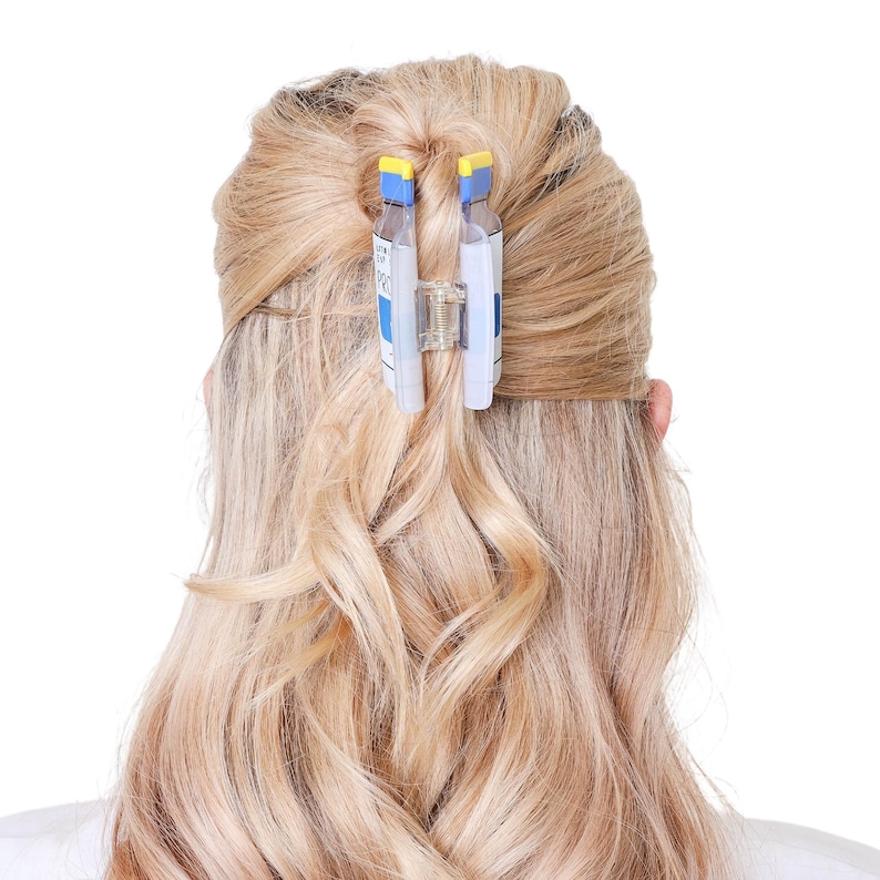 May include: A hair claw clip featuring a design that mimics a prescription medication bottle. The clip is clear with blue and white labels, yellow top accents, and a central metal spring mechanism, securing a section of blonde hair in place.