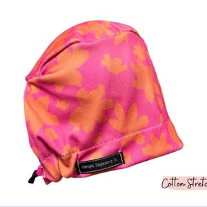 May include: A pink and orange floral patterned scrub cap. The cap is made of cotton stretch fabric and features a black tag with white text. The text "Cotton Stretch" is written in a cursive font.