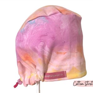 May include: A pink, yellow, and blue tie-dye patterned scrub cap. The cap has a drawstring and a small pink label. The words "Cotton Stretch" are printed on the bottom right corner.