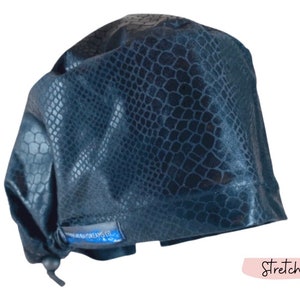 May include: A dark navy blue scrub cap featuring a glossy, embossed snakeskin texture pattern. The cap includes a gathered back with an adjustable toggle closure and a small blue rectangular fabric tag on the side with white text.