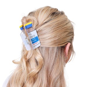 May include: A hair claw clip featuring a design shaped like a medical vial with a yellow cap. The label on the vial reads PROPOFOL 200mg/20mL with CAUTION printed in red. The clip is made of clear plastic and secures blonde hair in a half-up style.