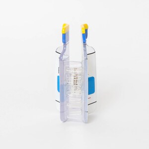 May include: A clear plastic hair claw clip featuring a white rectangular body with blue and yellow accents at the top. The clip has a central metal spring mechanism and interlocking teeth designed to hold hair securely in place.