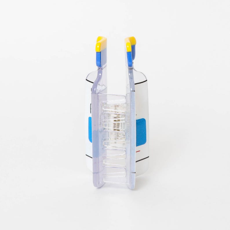 May include: A clear plastic hair claw clip featuring a white rectangular body with blue and yellow accents at the top. The clip has a central metal spring mechanism and interlocking teeth designed to hold hair securely in place.