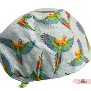 May include: A light blue cotton scrub cap featuring a repeating pattern of colorful parrots with outstretched wings. The parrots have yellow, orange, green, and blue plumage. The cap has a green drawstring and the word "Cotton" is printed on the image.