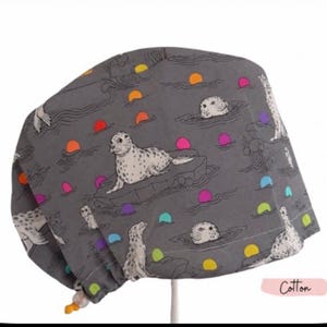 May include: A gray scrub cap featuring a repeating pattern of white spotted seals on ice floes and in water, accented by small, vibrant circles in orange, pink, yellow, and teal. The fabric is labeled as cotton in the bottom right corner.