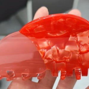 May include: A two-toned hair claw clip. The top half is a vibrant red with a marbled effect, while the bottom half is a translucent pink. The clip has a curved shape with teeth for securing hair. A stylish hair accessory.