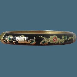 Op de afbeelding: Een zwarte scharnierende armband met een goudkleurige metalen afwerking en een ingelegd bloemmotief. Het ontwerp toont delicate bloemen met witte, rode en groene emaille accenten die rond de buitenkant van de cirkelvormige band lopen.
