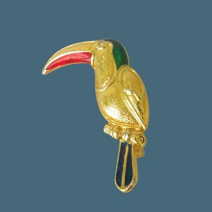 Op de afbeelding: Een goudkleurige toekan vogelbroche met een rode snavel, groene kop en zwart-gouden staart. De vogel zit op een tak. De broche is waarschijnlijk gemaakt van metaal en email.