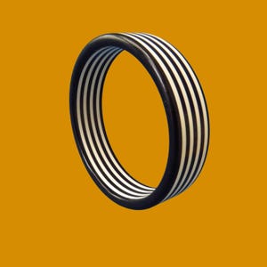 Pode incluir: Uma pulseira de bracelete listrada em preto e branco. A pulseira apresenta listras pretas e brancas alternadas. A joia está sobre um fundo laranja sólido. A pulseira tem formato circular.