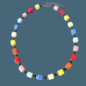 Op de afbeelding: Een kleurrijke ketting met vierkante kralen in verschillende kleuren, waaronder rood, geel, roze en blauw. De kralen zijn geregen met zwarte kralen en goudkleurige accenten, wat een levendig accessoire creëert. De ketting heeft een ketting sluiting.