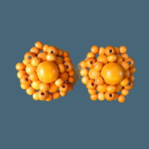 Puede incluir: Dos pendientes redondos de madera. Cada pendiente está compuesto por muchas cuentas pequeñas de madera de color naranja claro que rodean una cuenta central más grande del mismo color. Los pendientes tienen un diseño floral o de sol.
