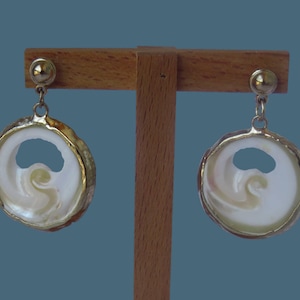 Op de afbeelding: Een paar ronde oorbellen met witte iriserende schelpkernen met natuurlijke spiraalpatronen. Elke schelp is gevat in een metalen goudkleurige rand en hangt aan een kleine goudkleurige stud met een gladde, gepolijste bolvormige bovenkant.