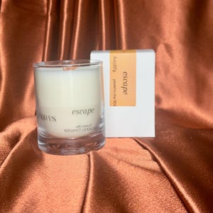 May include: A clear glass candle with a wooden wick and white wax, next to a white box. The candle and box both have the word "escape" printed on them. The candle is 8 oz.