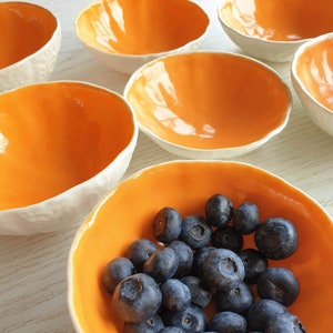 May include: A set of six small, white ceramic bowls with orange interiors. The bowls are arranged in a semi-circle, with one bowl in the centre filled with blueberries.