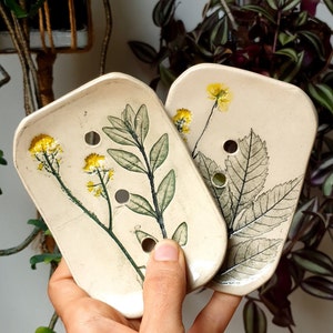 Peut inclure: Deux savonnettes en céramique avec des motifs floraux. Les savonnettes sont blanches avec des feuilles vertes et des fleurs jaunes. Chaque savonnette a trois trous de drainage.