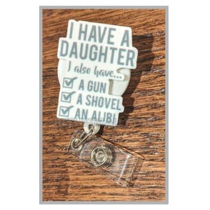 May include: A white badge reel with the text "I HAVE A DAUGHTER, I also have... A GUN, A SHOVEL, AN ALIBI" in gray lettering. The badge reel has a clear plastic retractable cord and a metal clip.
