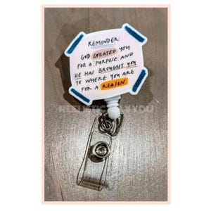 May include: White plastic badge holder with a blue and pink design. The badge holder has a retractable cord and a clip. The text on the badge holder reads: "Reminder: God created you for a purpose and he has brought you to where you are for a reason."