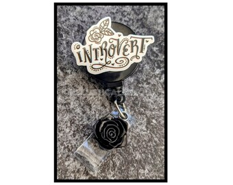 May include: Black retractable badge holder with a white sticker that says "Introvert" with a black rose charm.