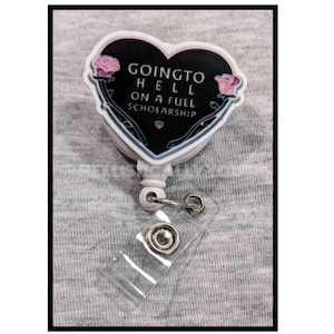 May include: White heart-shaped badge holder with a black and white design featuring the text "Going to Hell on a Full Scholarship" and pink roses. The badge holder has a clear plastic retractable reel.