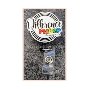May include: A white round badge holder with a black retractable cord and a clear plastic badge holder. The badge holder has a colorful graphic that says "Difference Maker #Teacherlife".
