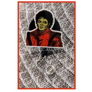 May include: A black and white retractable badge holder with a round, colorful image of Michael Jackson wearing a red jacket. The image is on a white background.