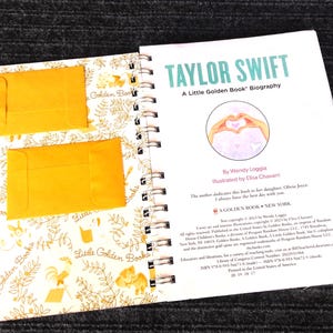 May include: An open book with a spiral binding, featuring the title "TAYLOR SWIFT" in teal. The book is a "Little Golden Book" biography, with two small yellow envelopes on the left page. The right page has a heart-shaped illustration.