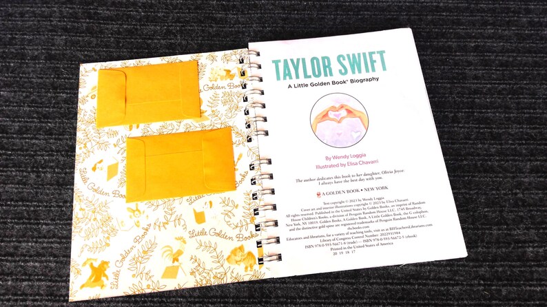 May include: An open book with a spiral binding, featuring the title "TAYLOR SWIFT" in teal. The book is a "Little Golden Book" biography, with two small yellow envelopes on the left page. The right page has a heart-shaped illustration.
