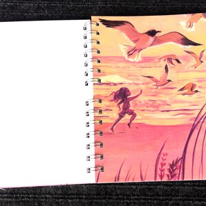 May include: Spiral-bound notebook with a colorful illustration of a person running on a beach with seagulls flying overhead. The artwork features a pink and orange sunset, creating a vibrant scene. The left side of the notebook is blank.