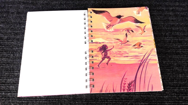 May include: Spiral-bound notebook with a colorful illustration of a person running on a beach with seagulls flying overhead. The artwork features a pink and orange sunset, creating a vibrant scene. The left side of the notebook is blank.
