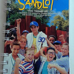 May include: A VHS tape cover for the movie "The Sandlot". The cover features a group of boys playing baseball, with the title of the movie in large yellow letters. The tagline reads "Two thumbs up! A charming and whimsical comedy about boys and baseball and a great big dog." - Siskel & Ebert