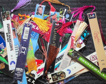 May include: A collection of colorful bookmarks with various movie and TV show themes, including "E.T.", "Ferris Bueller's Day Off", "Game of Thrones", and "Godzilla". Each bookmark features a tassel.