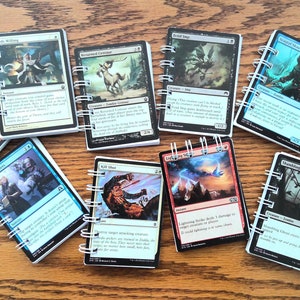 May include: A collection of Magic: The Gathering cards, each bound with a white spiral binding. The cards feature various fantasy illustrations and text, with a wooden surface in the background.