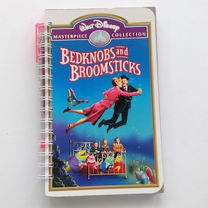 May include: A spiral-bound notebook featuring the Walt Disney Masterpiece Collection cover art for "Bedknobs and Broomsticks." The cover shows a couple dancing underwater, with children on a bed and colorful fish. A pink pen is attached.