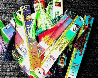 May include: A collection of colorful bookmarks made from recycled VHS tapes. The bookmarks feature various characters and titles, including "Frosty the Snowman", "Rudolph the Red-Nosed Reindeer", and "The Little Drummer Boy". Each bookmark has a tassel attached.