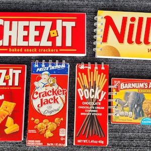 May include: A collection of six mini spiral notebooks featuring popular snack brands. The notebooks are red, yellow, and blue with images of the snacks and their logos. The brands include Cheez-It, Nilla Wafers, Cracker Jack, Pocky, and Barnum's Animals.