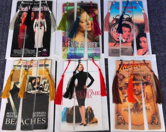 May include: A collection of movie-themed bookmarks. Each bookmark features a movie poster, such as "Death Becomes Her," "Ever After," "Groundhog Day," "Beaches," "Sweet Home Alabama," and "Indiana Jones." Each bookmark has a tassel.