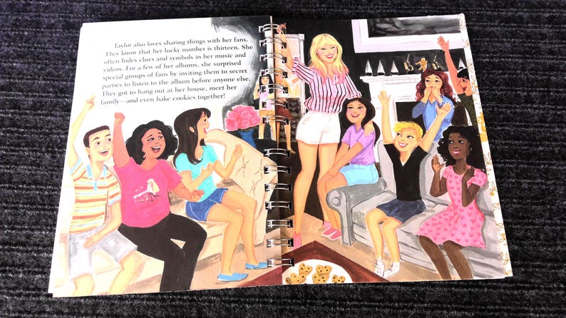May include: An illustrated book spread depicts a group of people, including a blonde woman in a striped shirt and white shorts, surrounded by other people. The text on the page describes sharing with fans.