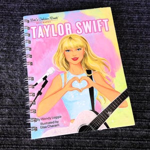 May include: A spiral-bound book titled "Taylor Swift Biography" from the Little Golden Book series. The cover features an illustration of Taylor Swift with blonde hair, forming a heart with her hands, and holding a guitar. The background is a mix of pink, yellow, and blue.