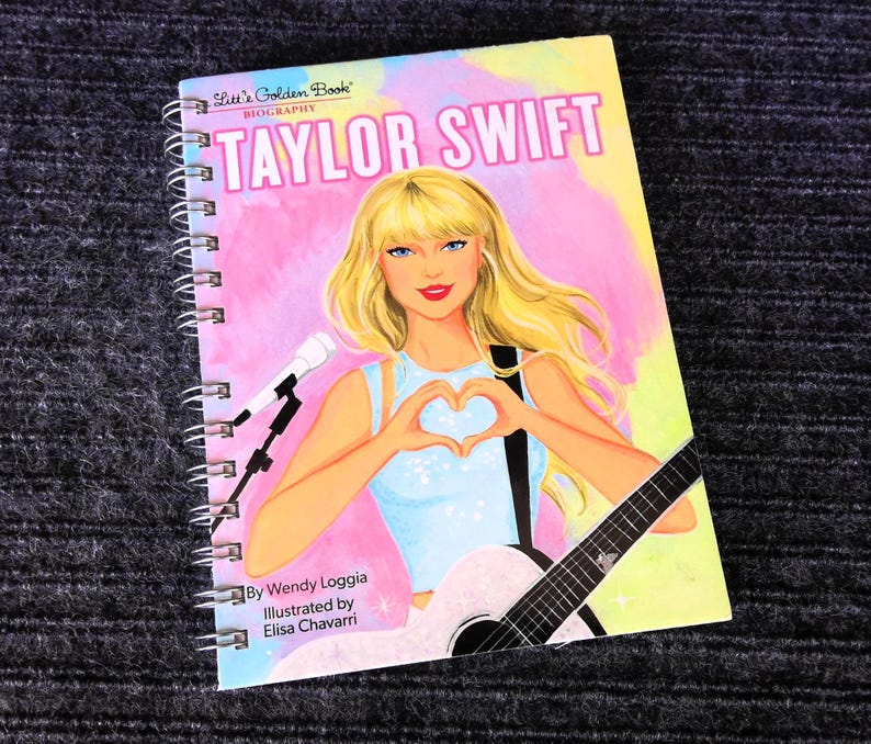 May include: A spiral-bound book titled "Taylor Swift Biography" from the Little Golden Book series. The cover features an illustration of Taylor Swift with blonde hair, forming a heart with her hands, and holding a guitar. The background is a mix of pink, yellow, and blue.