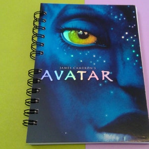 May include: A blue spiral notebook with a close-up image of a blue alien with a yellow eye. The notebook cover has the text "JAMES CAMERON'S AVATAR" in white.