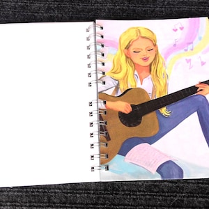 May include: Spiral-bound notebook with a drawing of a person playing a brown acoustic guitar. The person has long blonde hair, wearing a white shirt and blue jeans. Musical notes float in the background.