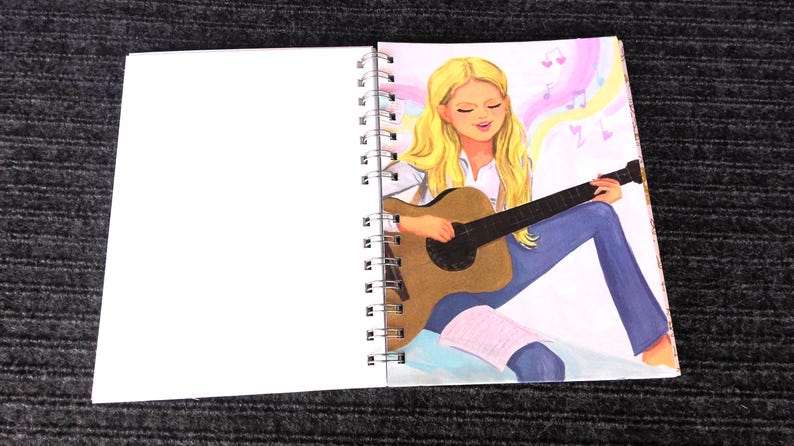 May include: Spiral-bound notebook with a drawing of a person playing a brown acoustic guitar. The person has long blonde hair, wearing a white shirt and blue jeans. Musical notes float in the background.