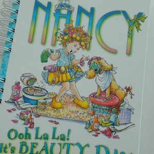 May include: A book cover featuring a cartoon girl with a flower crown, holding a hair dryer and a bottle of hairspray. She is standing next to a dog wearing a white bandana. The book title is "Fancy Nancy Ooh La La! It's Beauty Day" and the author and illustrator are Jane O'Connor and Robin Preiss Glasser.