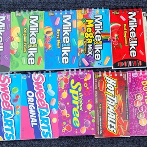 May include: A collection of 10 mini spiral notebooks featuring various candy brands, including Mike and Ike, Sweetarts, Spree, and Hot Tamales. The notebooks are colorful and feature the brand's logos and candy images.