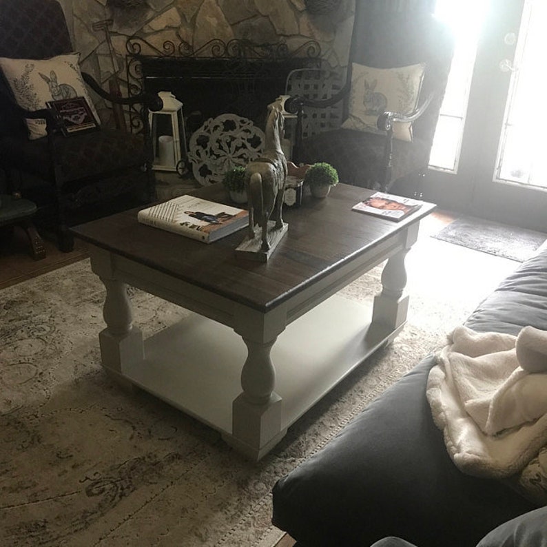 May include: A dark wood coffee table with a white painted base and a lower shelf. The table has turned legs and is sitting on a rug with a light brown and beige pattern.
