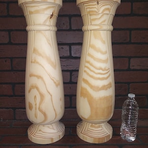May include: Two unfinished wooden balusters with a turned design. The balusters are approximately 18 inches tall and 4 inches in diameter. A wood chisel is lying on the floor in front of the balusters.