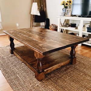 May include: A dark brown wooden coffee table with a lower shelf. The table has turned legs with a decorative design. The table is sitting on a woven rug.