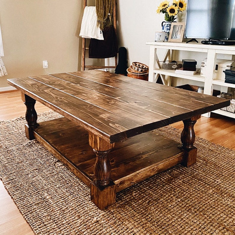 May include: A dark brown wooden coffee table with a lower shelf. The table has turned legs with a decorative design. The table is sitting on a woven rug.